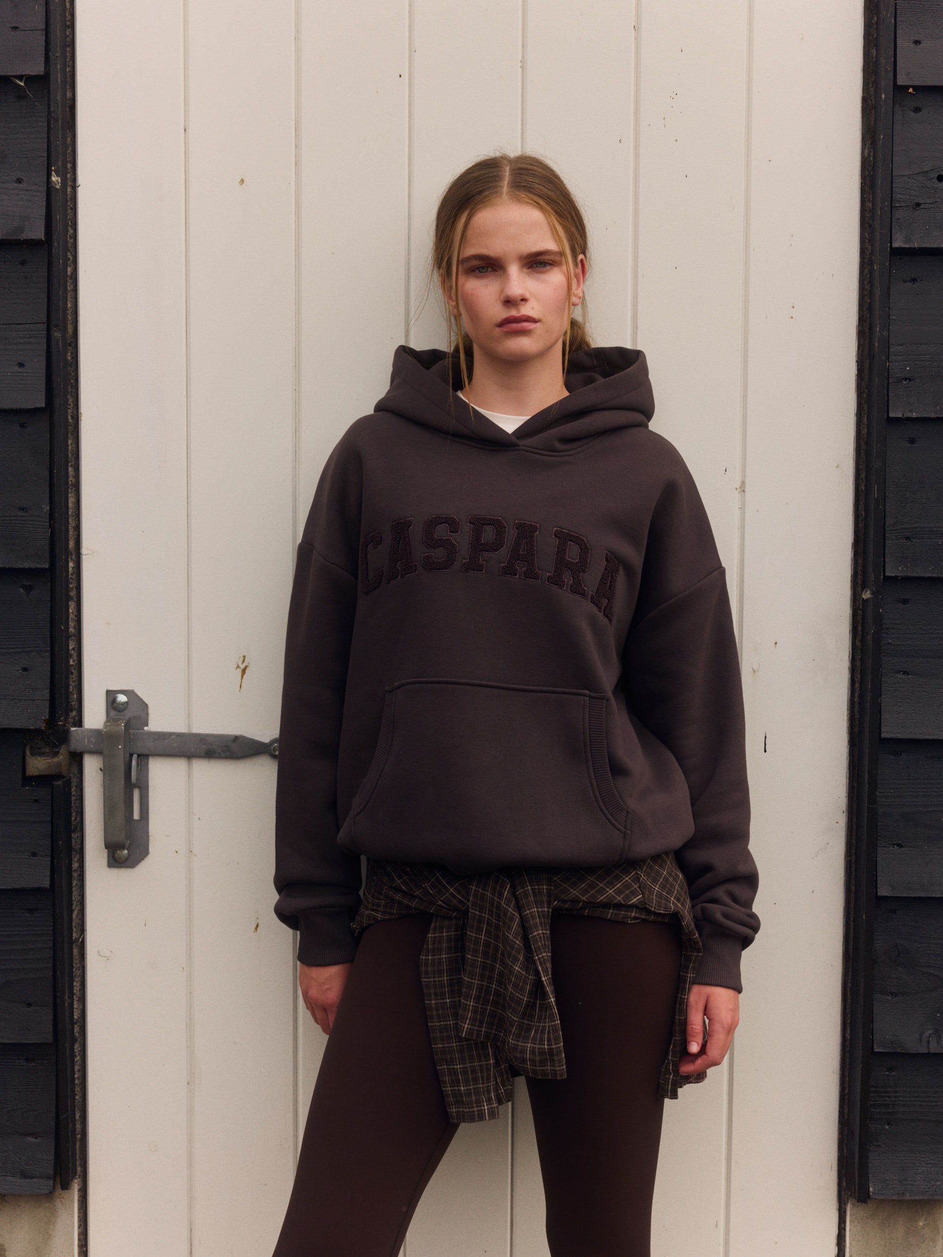 Woman wearing an espresso brown CASPARA hoodie standing in front of a white wooden door.